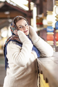 A slavic woman in a national colored scarf,a fur coat and  mittens on the porch of a wooden house