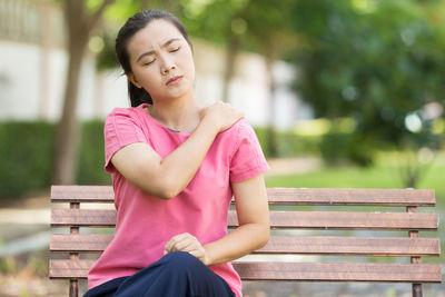 Young woman suffering from shoulder pain while sitting on bench at park