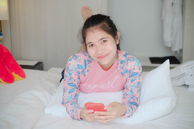Portrait of smiling young woman holding phone while relaxing on bed at home