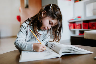 Focused girl drawing with pencil in album while sitting at wooden table in light room on blurred background at home