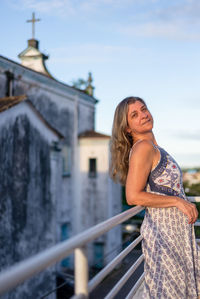 A woman on the porch of her house looking at the camera against the sky