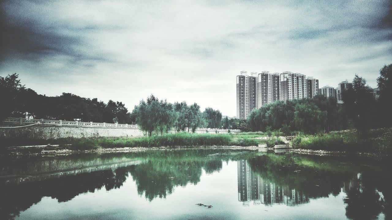 water, architecture, building exterior, built structure, tree, sky, reflection, city, tall - high, cloud, waterfront, scenics, cloud - sky, outdoors, skyscraper, modern, calm, tranquility, day, beauty in nature, tranquil scene, nature, cloudy, no people, office building, park