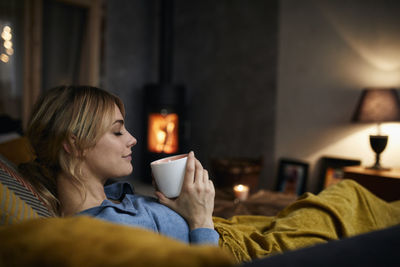 Smiling woman with cup of coffee relaxing on couch at home in the evening