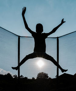Vertical front backlighting photo of a silhouette of a barefoot young boy jumping or flying on a trampoline with net and the sun at the background reflecting sunbeam rays on his shadow