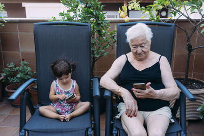 Grandmother and baby girl sitting side by side on the terrace using mobile phones