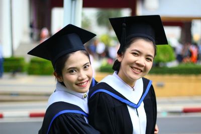 Portrait of smiling friends wearing graduation gown while standing outdoors