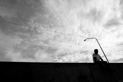 Low angle view of man sitting on railing against cloudy sky