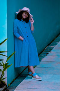 A young girl wearing a dress and a hat standing and leaning at a wall 