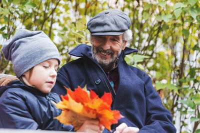 Happy senior man looking at great grandson holding maple leaves in park