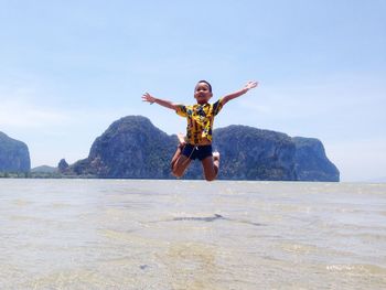 Portrait of young woman jumping on rock formation in sea against sky