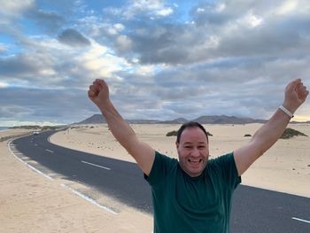 Full length of smiling man celebrating success while standing on sand at beach against sky