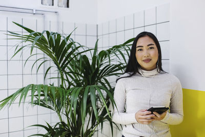 Portrait of smiling young woman holding smart phone while standing by plant in office