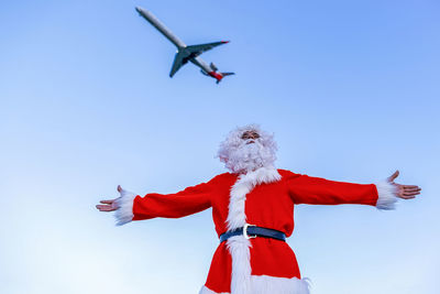 Man wearing santa claus costume standing with arms outstretched below flying airplane