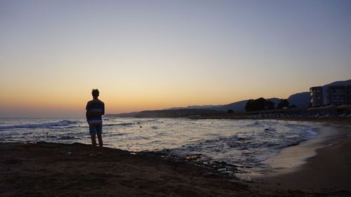 Rear view of man standing on beach against sky during sunset