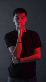 Portrait of young man with finger on lips standing against black background