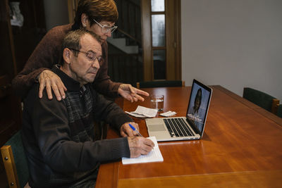 Senior man with social worker writing while video consultation with female doctor at home