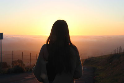 Rear view of woman wearing sweater standing on road against cloudscape during sunrise