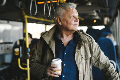 Businessman looking away holding disposable cup while standing in bus