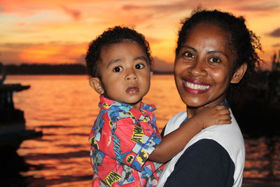 Portrait of smiling mother carrying son standing against sea during sunset