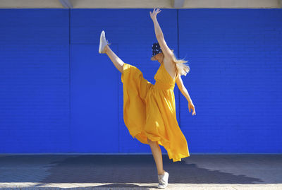 Carefree woman dancing by blue wall on sunny day