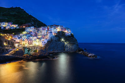 Illuminated buildings on rock by sea against blue sky