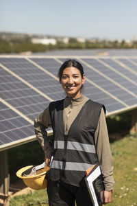 Portrait of confident female engineer standing with hand on hip near solar panels in field