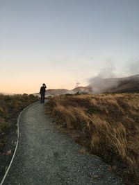 Full length of person standing on road amidst grassy field against clear sky during sunset