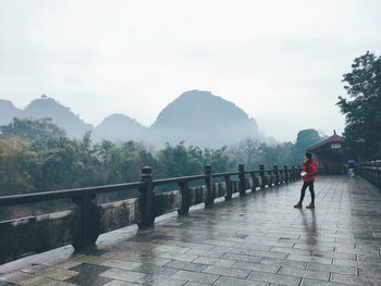 A girl standing on wet bridge looking at mountains in the mist