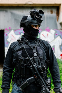 Serious young unrecognizable male warrior in modern protective wear balaclava and helmet with professional rifle looking away while standing near old building with graffiti on metal wall