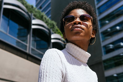 From below positive young african american woman in trendy outfit looking away and enjoying sunlight while sitting on bench on modern city street