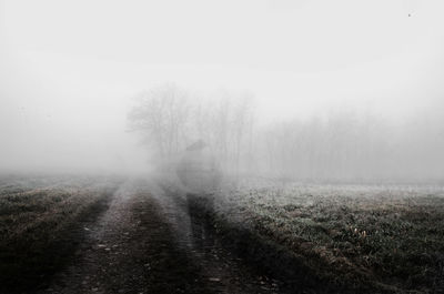 Dirt road amidst field against sky during foggy weather