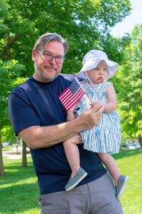 Portrait of grandfather carrying cute granddaughter with small american flag in park