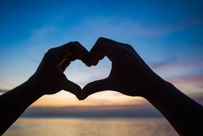 Close-up of hand holding heart shape against sea during sunset