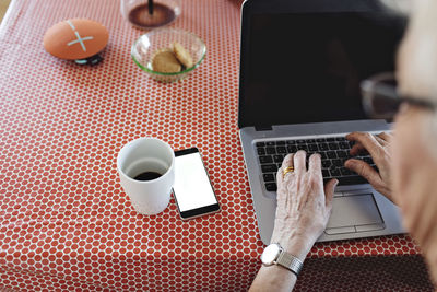 High angle view of senior woman using laptop by smart phone and coffee cup on dining table at home