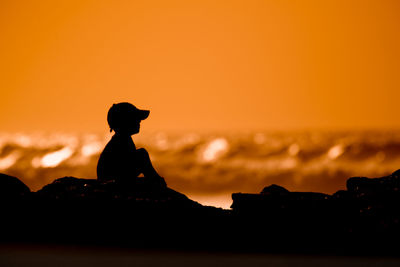 Silhouette boy sitting on rock at beach against clear sky during sunset