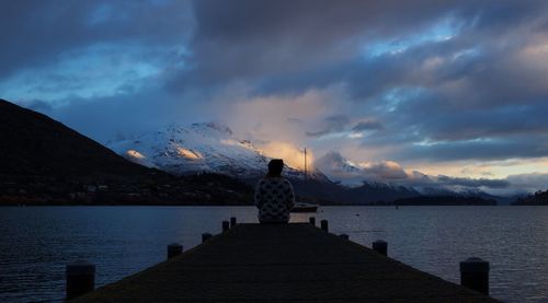 Rear view of woman sitting on pier at lake against sky at dusk
