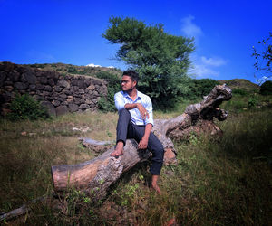 Full length of young man sitting on fallen tree over land against sky