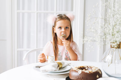 Funny cute little girl in light pink dress holding popcake in hands on festive table in living room 