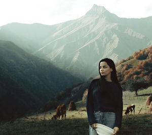 Young woman looking away while standing on grass against cows and mountains