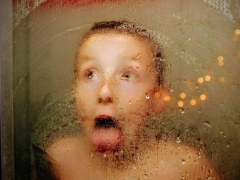 Cute boy sticking out tongue seen through wet condensed window in rainy season