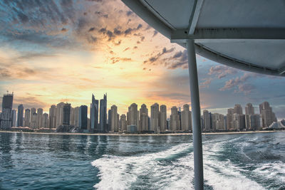 Sea by modern buildings against sky during sunset