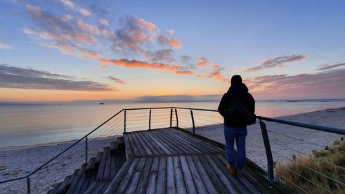 Rear view of man standing by sea against sky during sunset