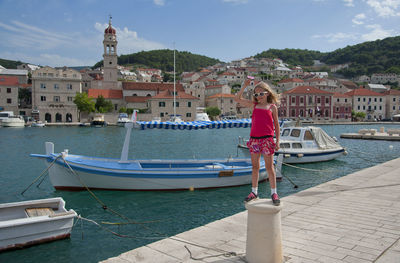 Full length of girl wearing sunglasses standing on bollard by river against buildings in city