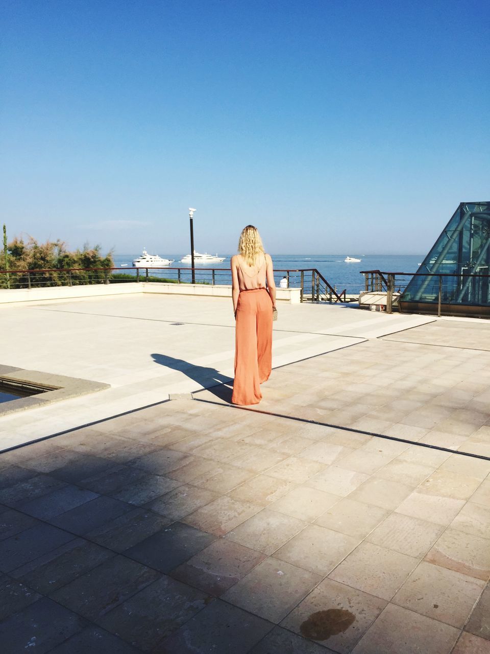 clear sky, copy space, water, rear view, blue, lifestyles, leisure activity, full length, standing, sea, casual clothing, sunlight, built structure, person, railing, architecture, day, outdoors