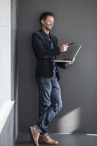 Happy businessman gesturing with pen while holding laptop against wall