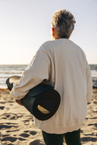 Rear view of senior woman looking at sea holding exercise mat while standing at beach