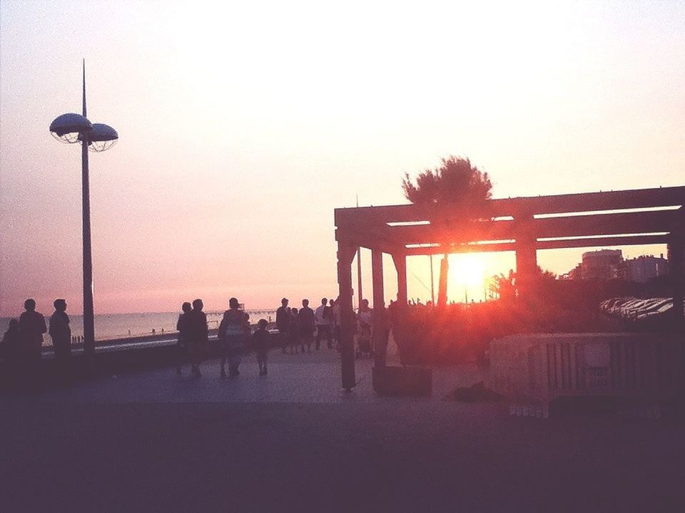 sunset, clear sky, sun, street light, large group of people, orange color, silhouette, built structure, copy space, architecture, sea, leisure activity, lifestyles, sunlight, water, beach, incidental people, sky, person