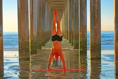 Shirtless man practicing yoga amidst architectural columns of pier at beach