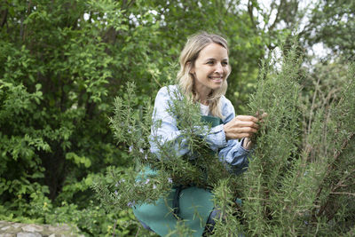 Young woman smiling while holding plants against trees