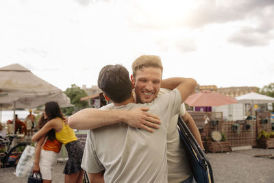 Happy young man embracing male friend with women hugging in background